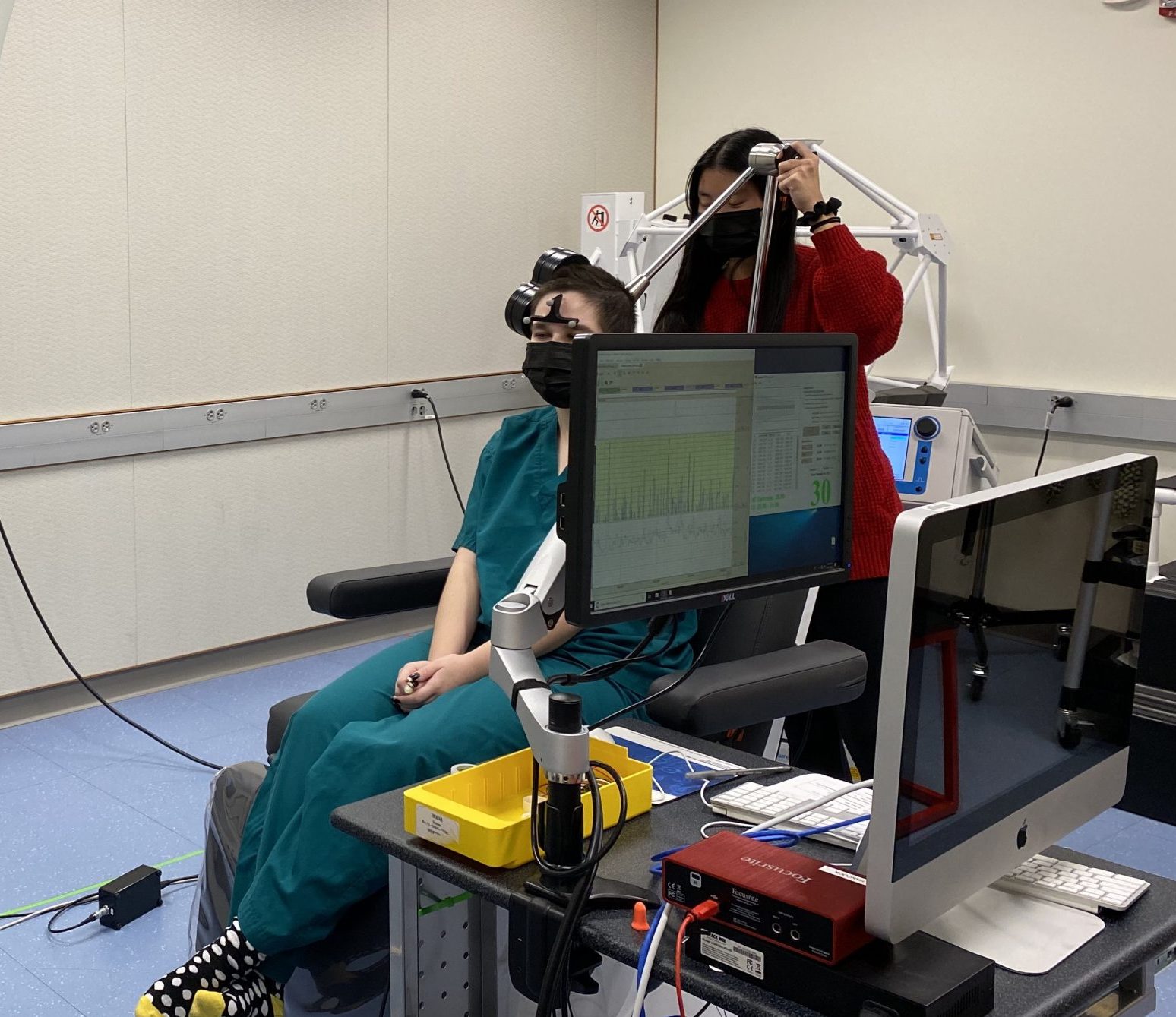 A demo setup of a transcranial magnetic stimulation (TMS) system. A participant sits in a chair with their back facing the camera and a TMS coil positioned over their head on the left.