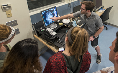 A researcher points at the MRI image of a brain on a screen to a group of high school students