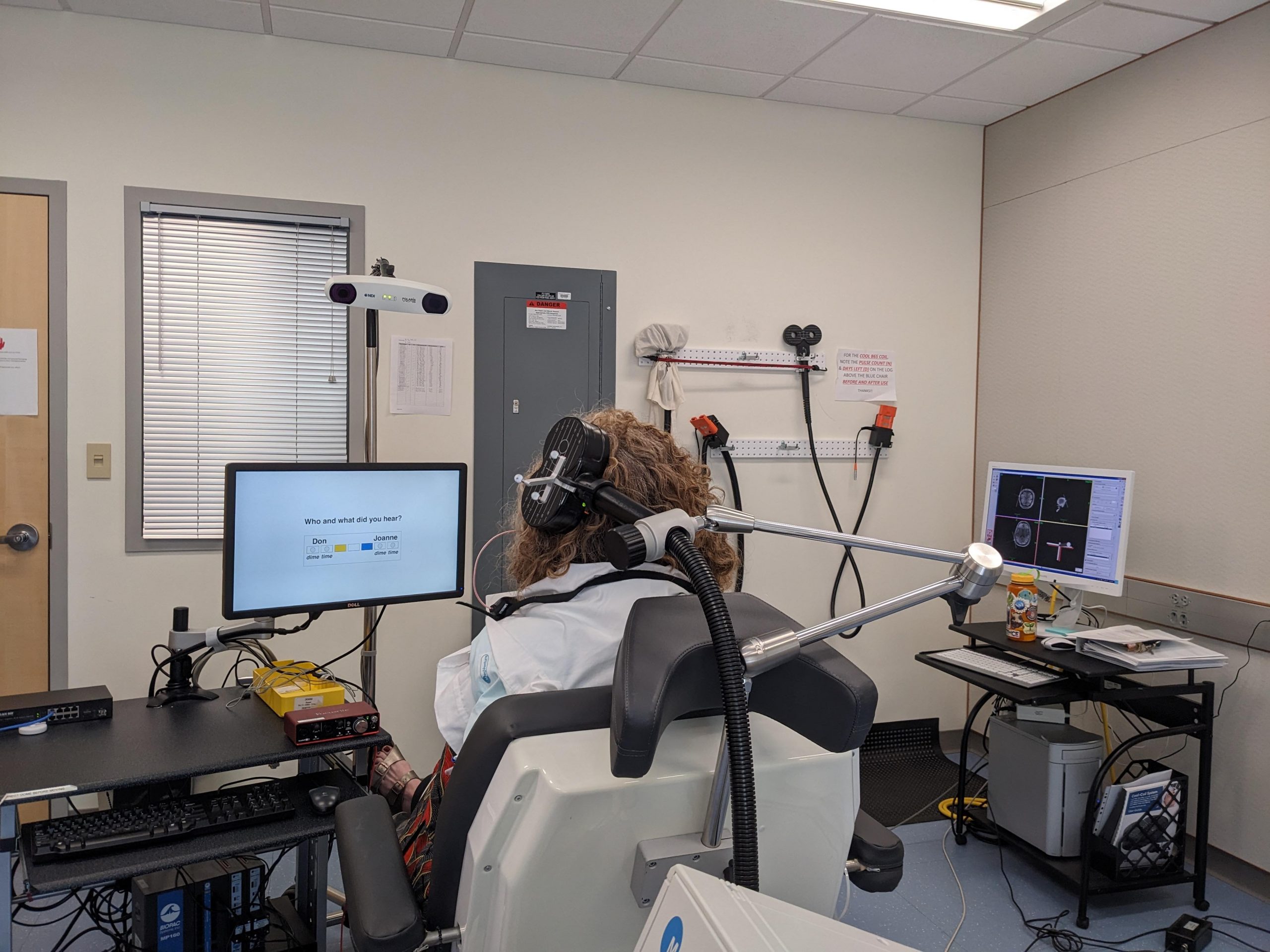 A demo setup of a transcranial magnetic stimulation (TMS) system. A participant sits in a chair with their back facing the camera and a TMS coil positioned over their head on the left.